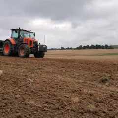 Pomarańczowy traktor z siewnikiem pracuje na polu, przygotowując ziemię do zasiewów. Dzierżawa ziemi w pełni.