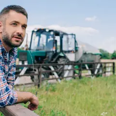 Rolnik w kratę oparty o płot, w tle traktor. Wsparcie dla małych gospodarstw to klucz do rozwoju wsi.