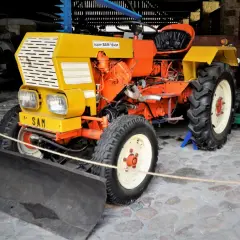 Pomarańczowo-żółty traktor "Super SAM 18 KM" z pługiem z przodu, gotowy do pracy.