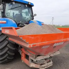 Niebieski traktor z rozsiewaczem napełnionym wapnem, gotowy do pracy na polu. Wapnowanie gleby poprawia jej strukturę i żyzność.