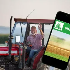 Uśmiechnięty rolnik w traktorze macha do kamery, obok smartfon z aplikacją eKRUS, która ułatwia zarządzanie prawem do emerytury rolniczej według nowych zasad.