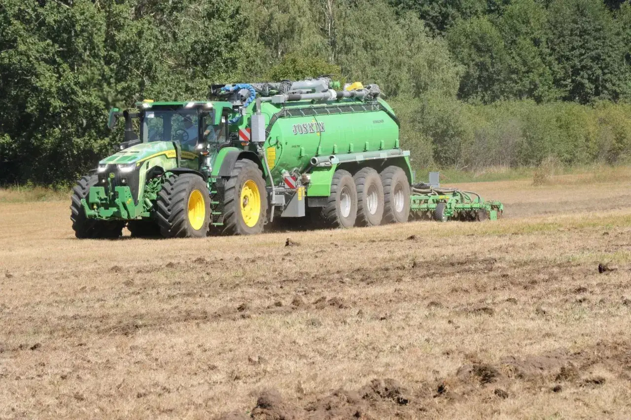 Zielony traktor z przyczepą asenizacyjną Joskin, wyposażony w system isobus, pracuje na polu, przygotowując glebę.