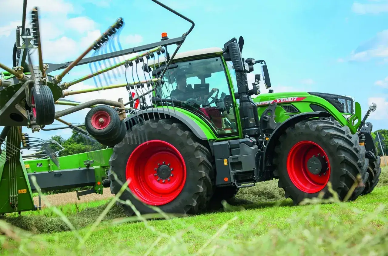 Zielony traktor Fendt 724 Vario z przyczepioną zgrabiarką pracuje na polu, zbierając siano.