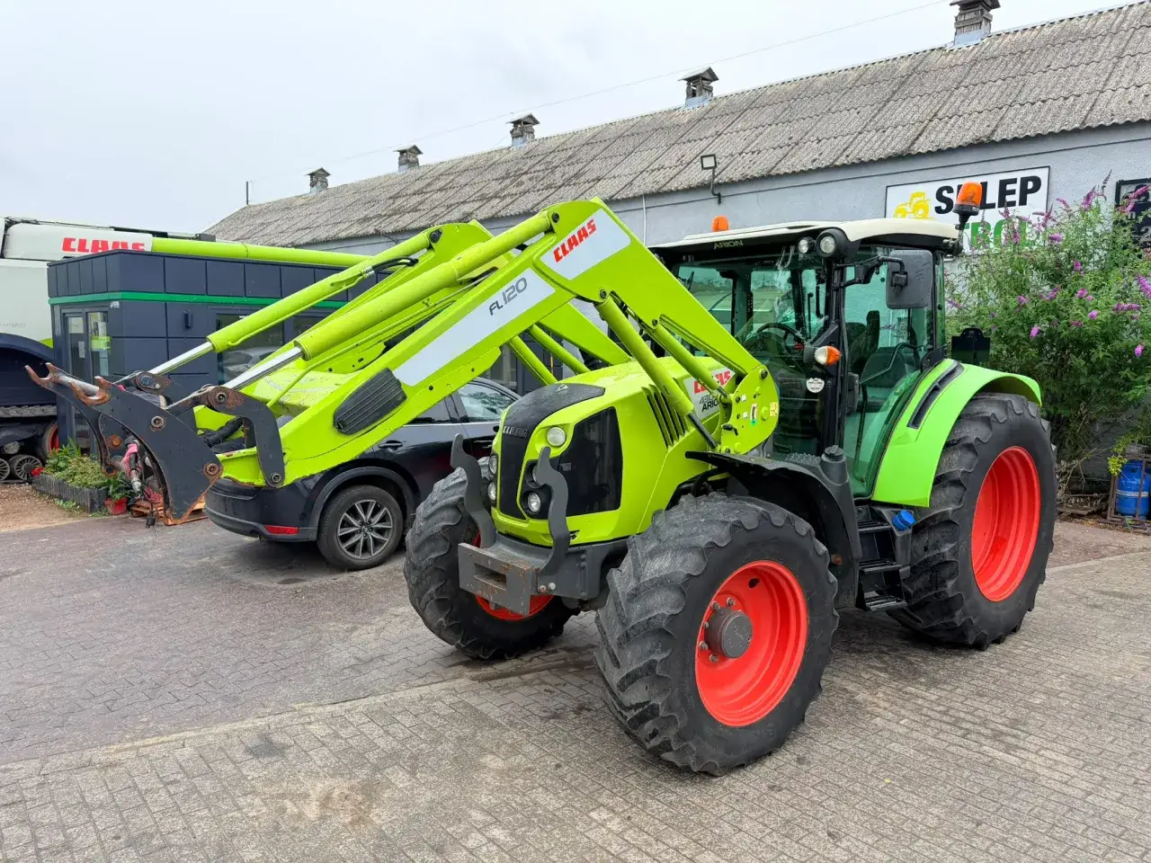 Zielony traktor Claas Arion 430 z ładowaczem czołowym FL120 stoi na placu przed budynkiem.