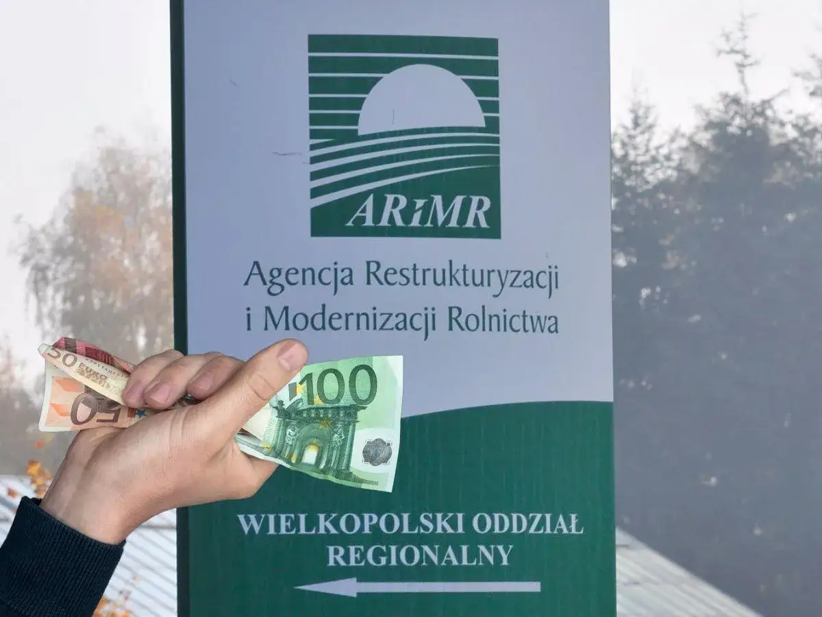 Ręka trzyma banknoty 50 i 100 euro przed tablicą ARiMR. Może to symbolizować arimr kredyt na zakup ziemi.